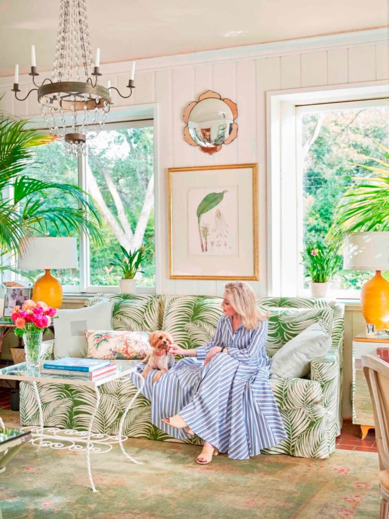A woman with a small white dog sits on a sofa in a palm leaf pattern